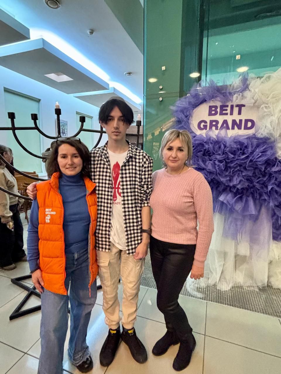 Дві жінки та юнак стоять разом у приміщенні біля входу з написом: BEIT GRAND, прикрашеним фіолетовими та білими декораціями, а поруч стоїть велика Менора.