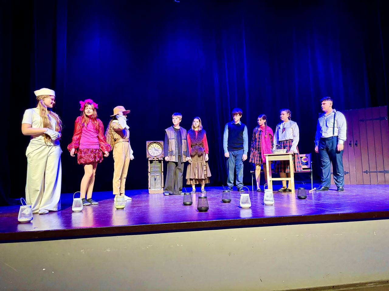 На сцені театру, освітленій інтенсивним синім світлом, стоять  дев'ять молодих акторів (діти та підлітки) у різних костюмах. Фон сцени повністю чорний, що підкреслює фігури акторів. На передньому плані сцени, біля ніг акторів, розміщено кілька маленьких ліхтариків. На сцені, праворуч, стоїть низький дерев'яний стіл.