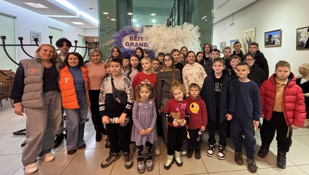 Групова фотографія дітей, підлітків та дорослих, які стоять разом у холлі театру. На задньому плані видно велику декоративну інсталяцію з білого та фіолетового пухнастого матеріалу з табличкою в центрі: Beit Grand. На стінах праворуч висять картини. Люди одягнені у теплий повсякденний одяг, посміхаються та позують.