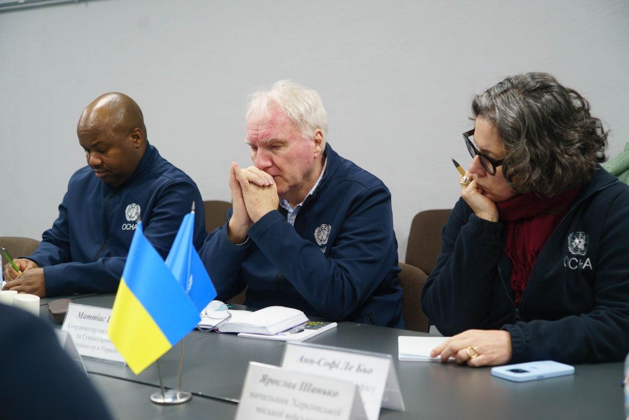 Троє представників міжнародної організації (OCHA) у фірмових темно-синіх кофтах сидять за робочим столом під час офіційної зустрічі. Перед ними встановлено маленькі прапорці України та ООН.