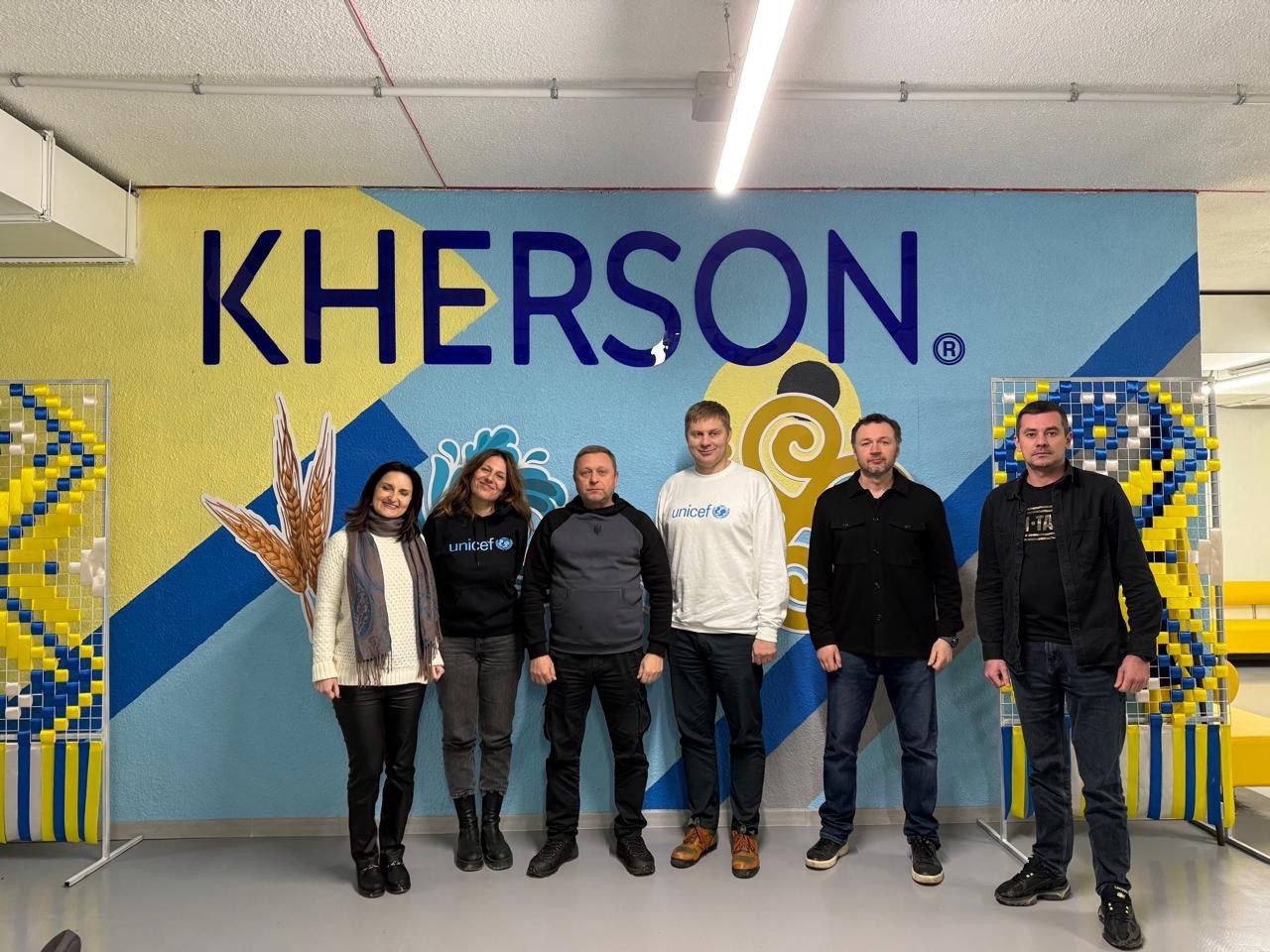 Група з шести осіб стоїть на фоні яскравої стіни з великим написом: KHERSON, виконаним у синьо-жовтих кольорах із патріотичним орнаментом. Двоє учасників одягнені у брендований одяг UNICEF, а з боків розміщені декоративні панелі з національною символікою.