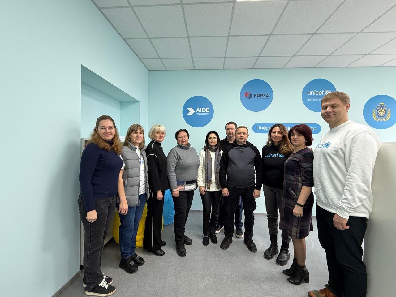 Групова фотографія представників UNICEF та партнерських організацій у світлому кабінеті цифрового освітнього центру. На блакитній стіні розміщені логотипи партнерів, зокрема UNICEF, AIDE та Республіки Корея. Учасники зустрічі посміхаються, позуючи на фоні облаштованого навчального простору.