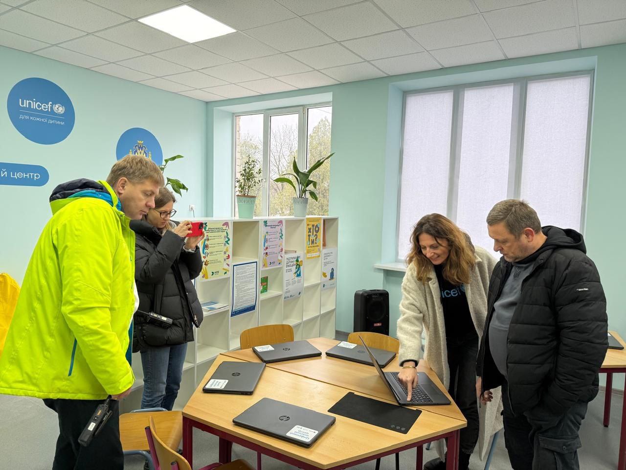 Представники влади та міжнародних організацій оглядають технічне оснащення цифрового освітнього центру. Жінка в брендованому одязі UNICEF демонструє роботу ноутбука HP на столі, навколо якого зібралися колеги у світлому кабінеті з великими вікнами та логотипами партнерів на стінах.