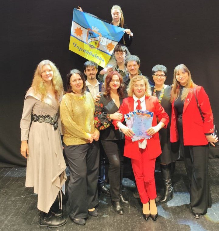 Група з десяти людей стоїть на сцені, позуючи для фото. Більшість одягнені у сучасний одяг. Жінка в центрі, одягнена в червоний костюм, тримає сертифікати або нагороди. На задньому плані інша жінка тримає прапор Чорнобаївки.