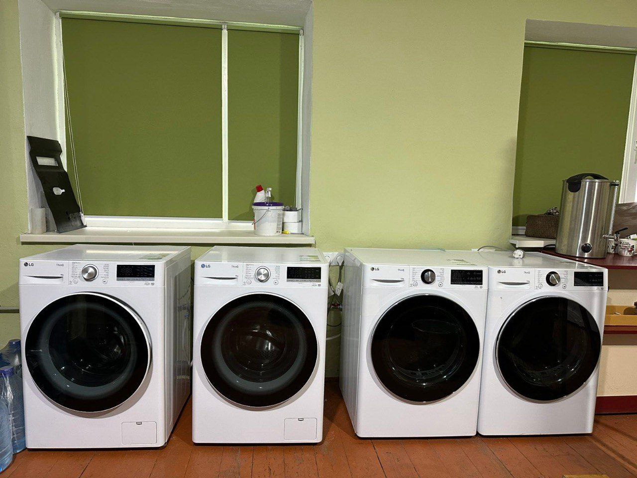 Чотири білі пральні машини LG стоять в ряд уздовж салатової стіни під вікном.
