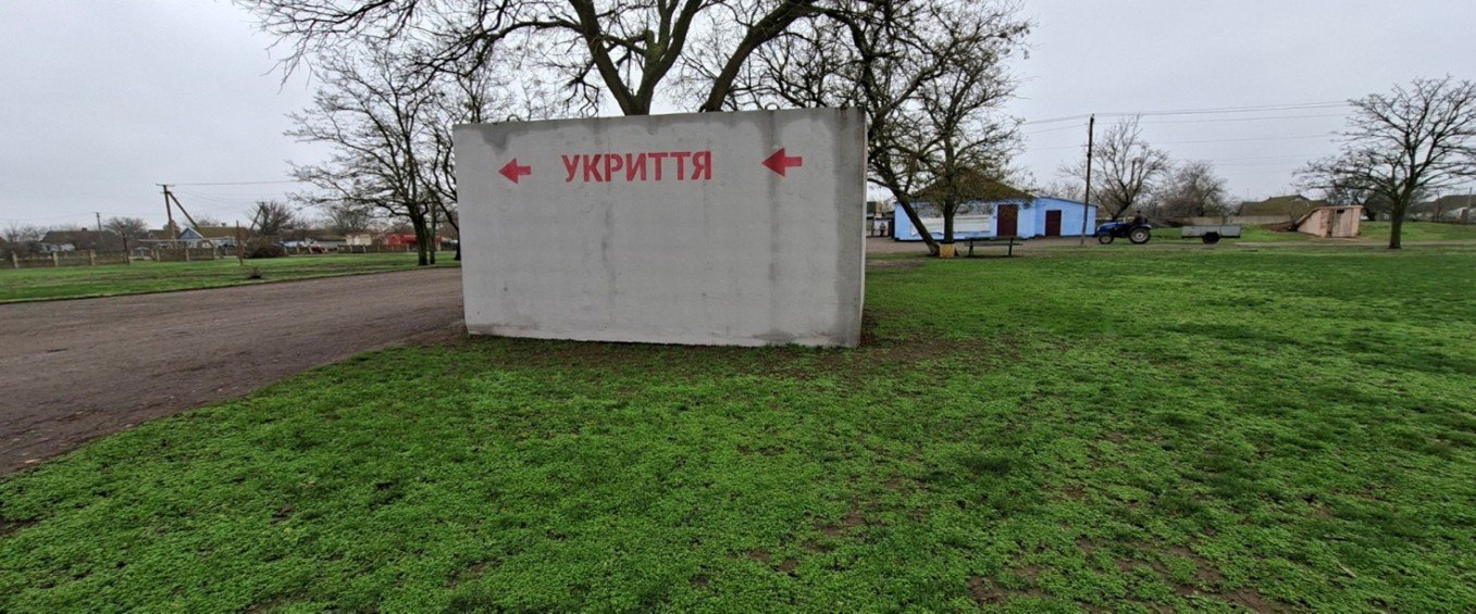 Бетонна споруда із написом червоними літерами: УКРИТТЯ та двома стрілками, що вказують у протилежні боки, стоїть на зеленій галявині в сільській місцевості.