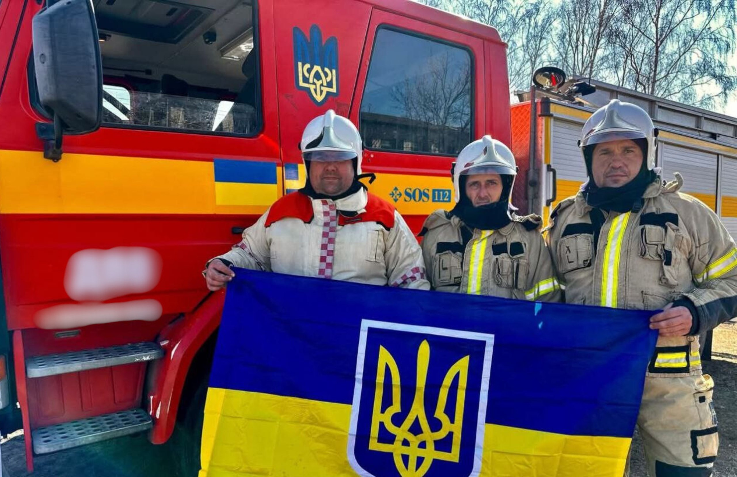 Троє рятувальників у повному екіпіруванні та шоломах стоять перед червоною пожежною машиною, тримаючи великий український прапор із Тризубом.