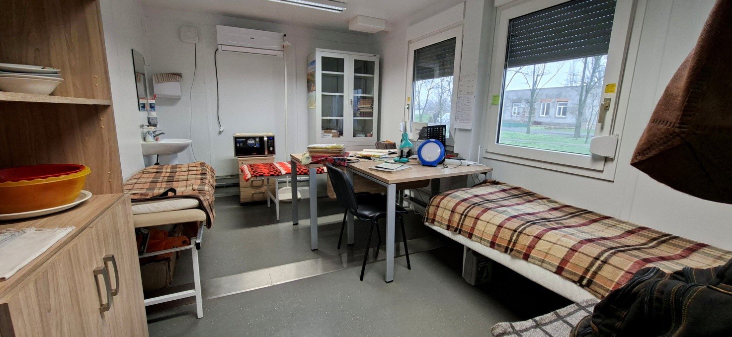 Інтер'єр лікарського кабінету з двома ліжками, застеленими картатими ковдрами, письмовим столом, шафою та великими вікнами.