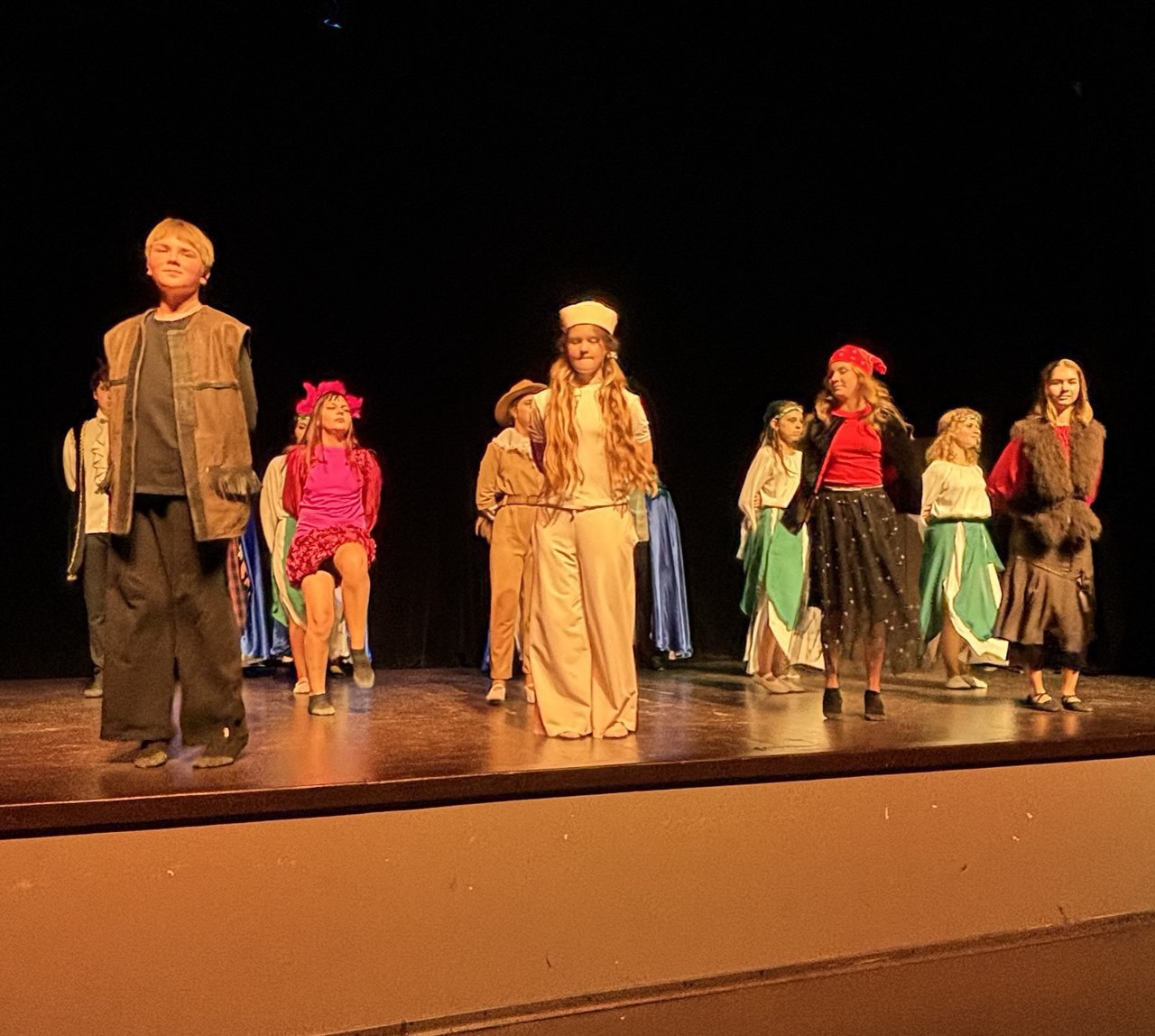 Група підлітків у різноманітних сценічних костюмах, схожих на персонажів вистави, стоїть на темній сцені, освітленій теплим світлом.