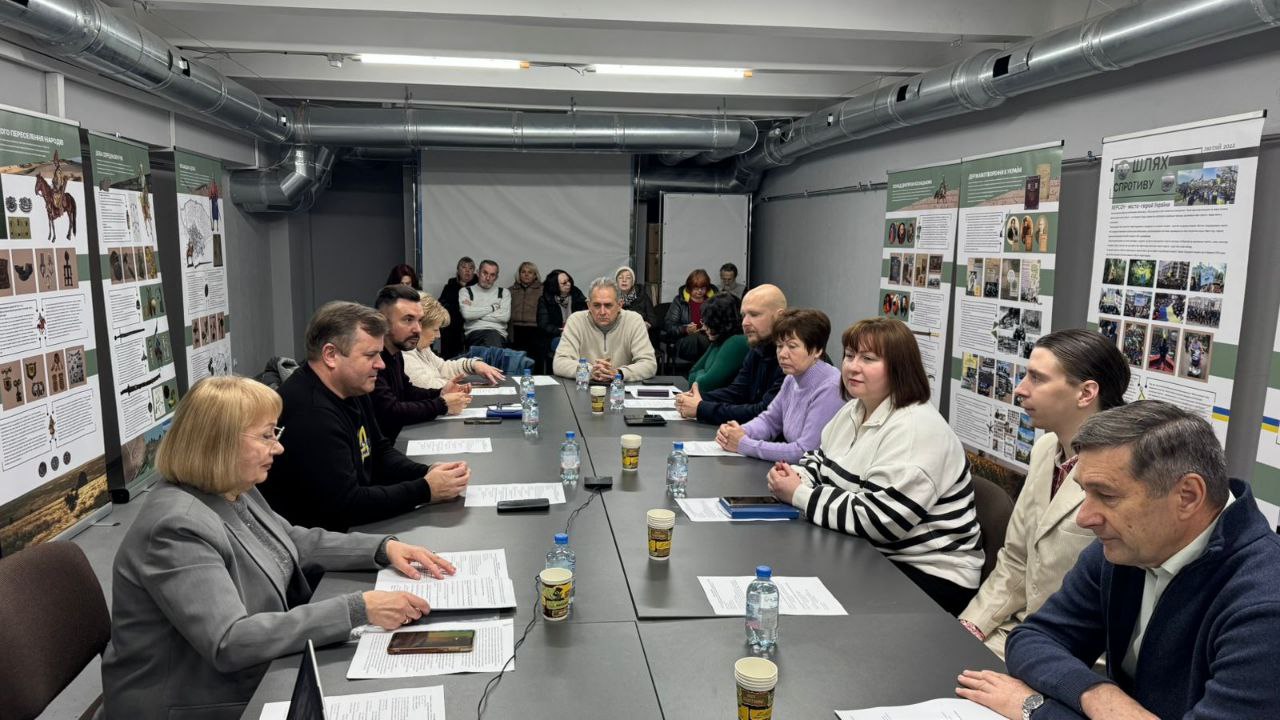 Група людей проводить зустріч за довгим столом у сучасному залі зі світло-сірими стінами та видимими вентиляційними трубами; уздовж стін розміщені інформаційні стенди про історію та спротив Херсонщини.