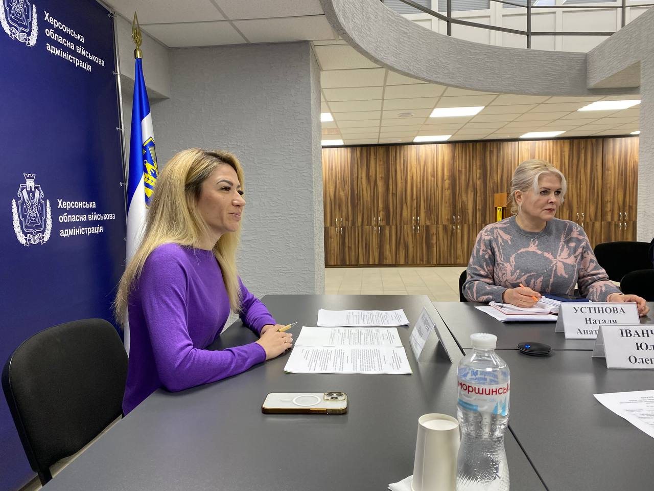 Нарада в офісному приміщенні: за столом сидять дві жінки, перед ними лежать документи; на фоні — прапор України та банер Херсонської обласної військової адміністрації.