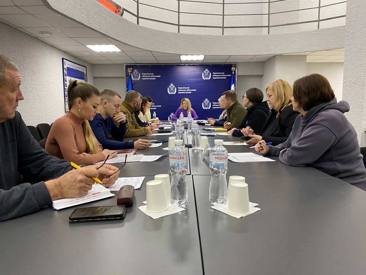 Група людей проводить робочу нараду за довгим столом у світлому офісному приміщенні на фоні банера Херсонської обласної військової адміністрації.