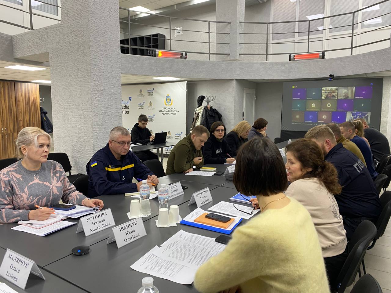 Робоча нарада у просторому залі з круговим балконом: учасники сидять за столом із документами, на фоні — банер Офіційний медіацентр. Херсонська обласна військова адміністрація та екран із відеозв'язком.