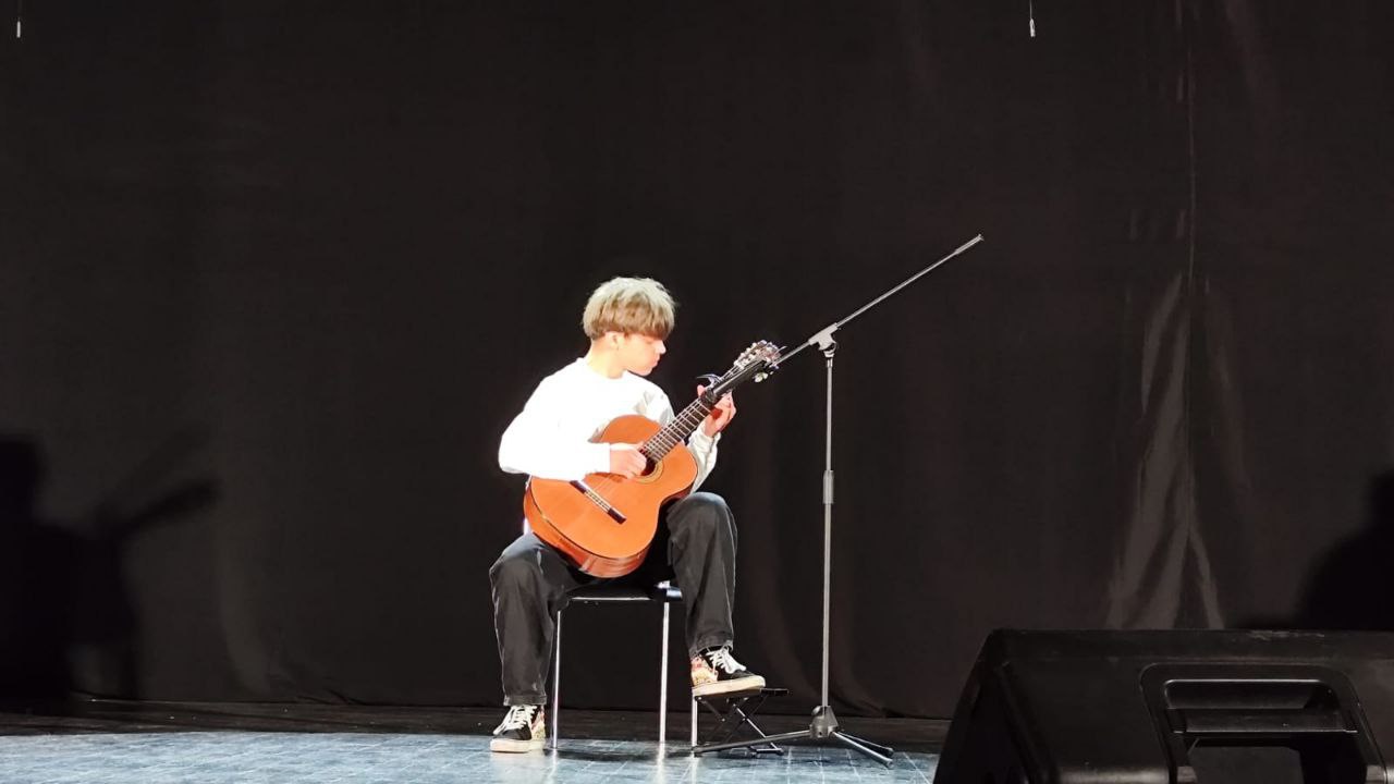 Молодий чоловік сидить на стільці на сцені на тлі чорної завіси, граючи на акустичній гітарі. Він одягнений у білий светр і чорні джинси.