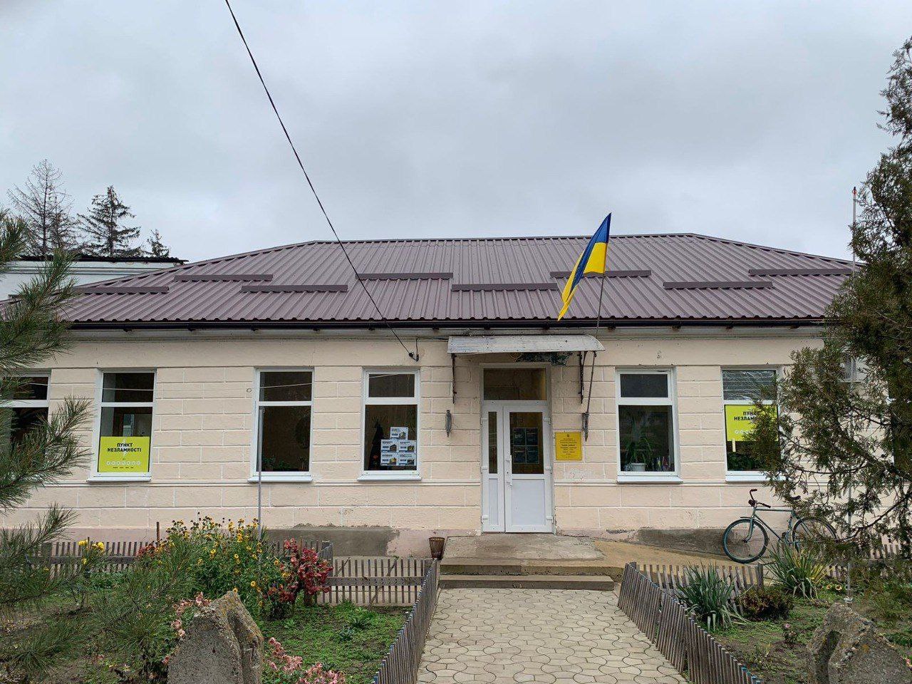 Світло-рожева будівля з коричневим дахом, білими вікнами та табличкою «Пункт незламності». Перед входом — доріжка з бруківки, невеликий паркан та квіти. На фасаді встановлено державний прапор України.