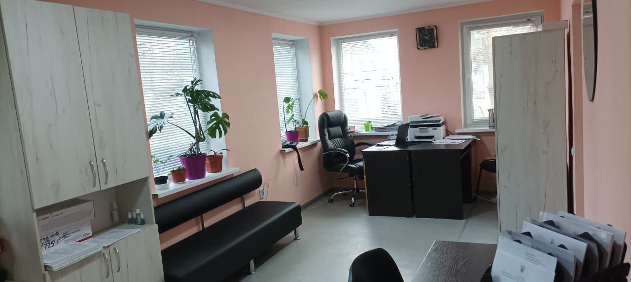 Світле офісне приміщення з рожевими стінами, робочим столом, чорним шкіряним кріслом, диваном та кімнатними рослинами на підвіконнях.