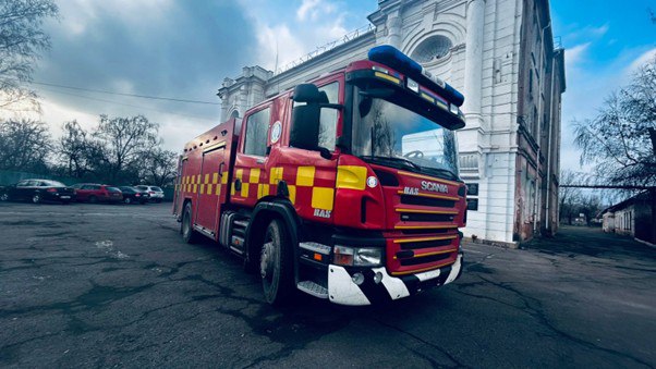Червоний пожежний автомобіль Scania з жовтим світловідбивним маркуванням, припаркований на асфальтованому майданчику на фоні великої білої будівлі.