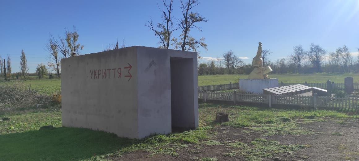 Бетонне наземне укриття прямокутної форми на відкритій місцевості. На бічній стіні червоною фарбою зроблено напис: УКРИТТЯ зі стрілкою, що вказує на вхід. Позаду розташований пам'ятник та зелені насадження під ясним небом.