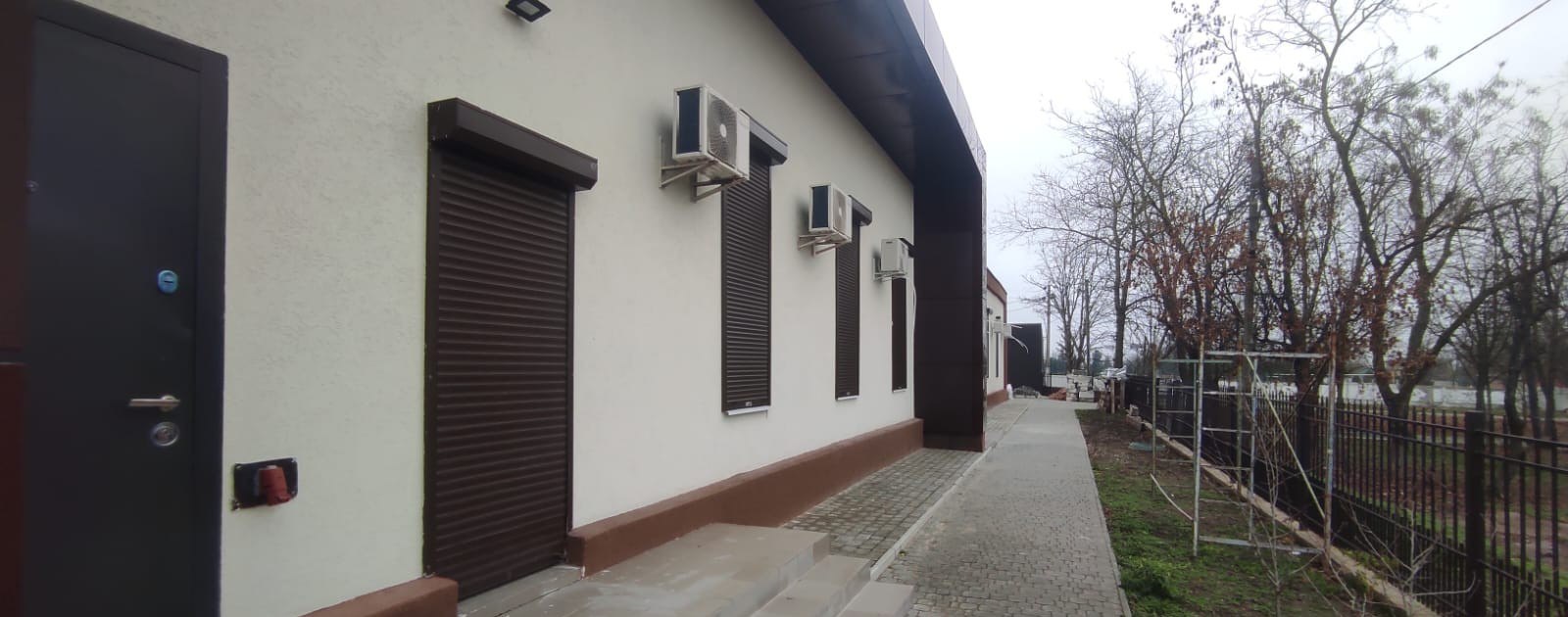 Вигляд збоку на сучасну будівлю з декількома вікнами, закритими коричневими ролетами, та встановленими кондиціонерами на світлій стіні.