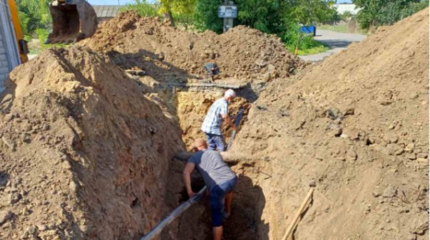Двоє чоловіків прокладають підземні комунікації на відкритій місцевості серед куп викопаного ґрунту у глибокій траншеї за допомогою екскаватора.