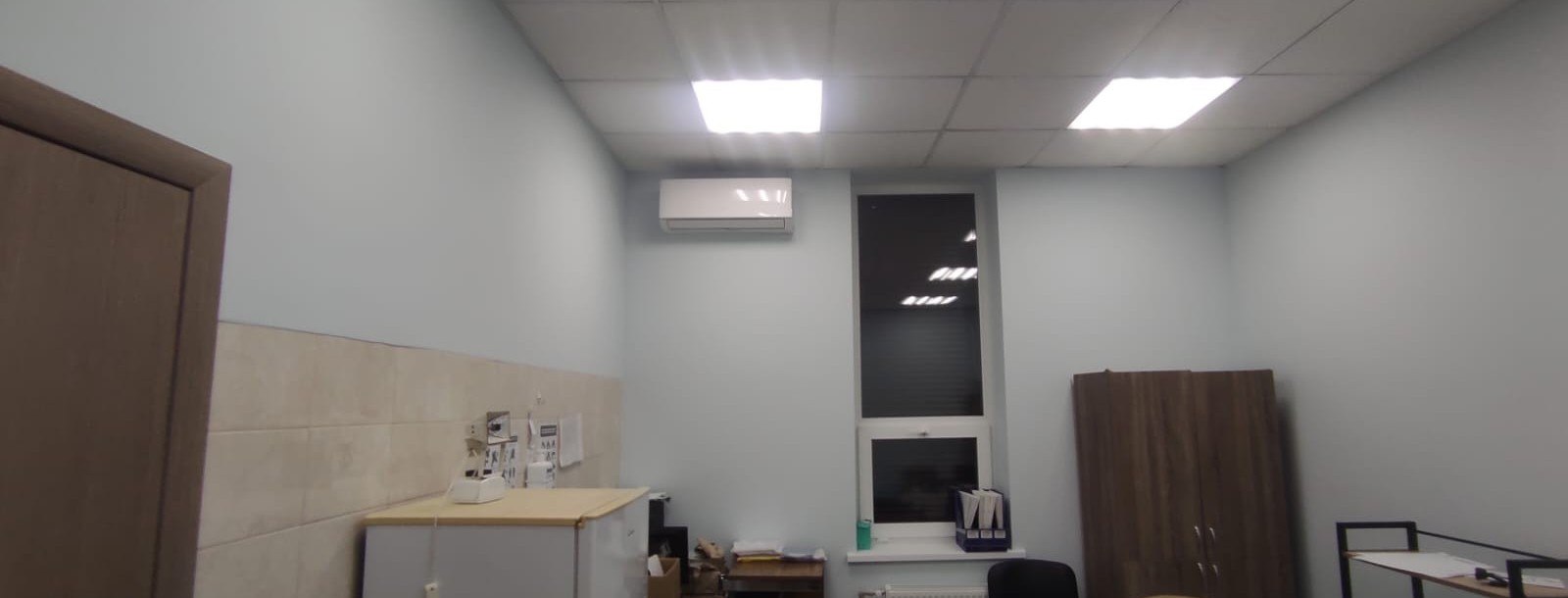 Інтер'єр робочого кабінету зі світло-блакитними стінами, офісними меблями, вікном та кондиціонером під стелею.