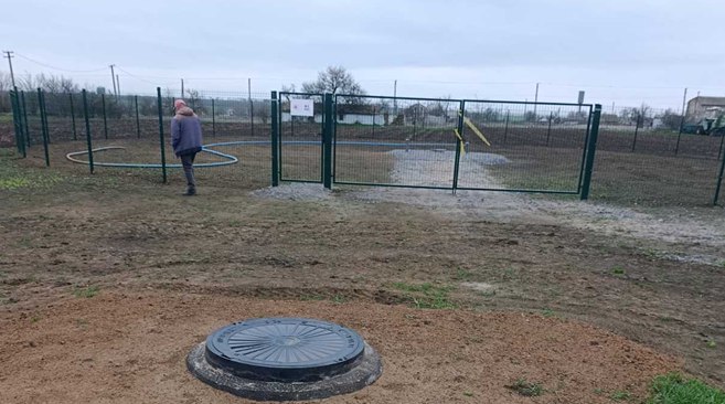 Огороджена сітчастим парканом територія з люком на передньому плані, технічними шлангами на землі та людиною, яка йде вздовж паркану.