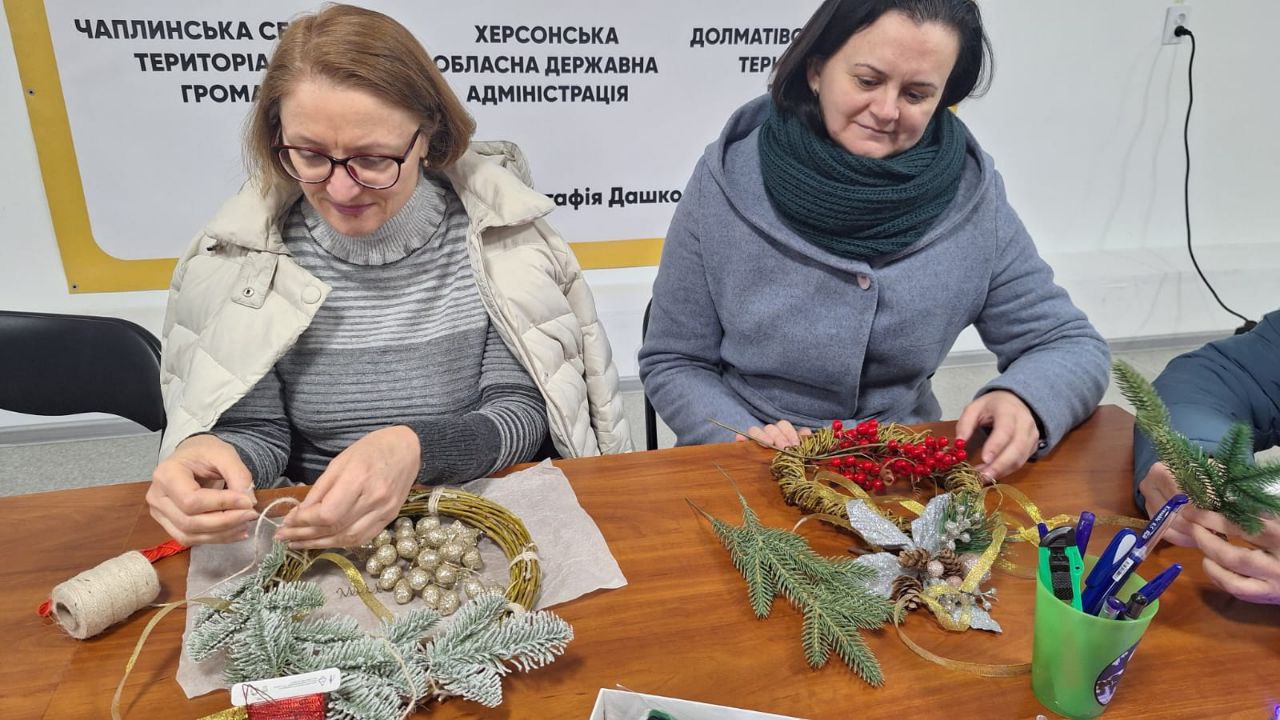 Двоє учасниць заходу зосереджено майструють святкові вінки, використовуючи золотисте листя, хвойні гілки, червоні ягоди та шпагат на фоні банера Чаплинської громади, Херсонської ОДА та Долматівської територіальної громади.