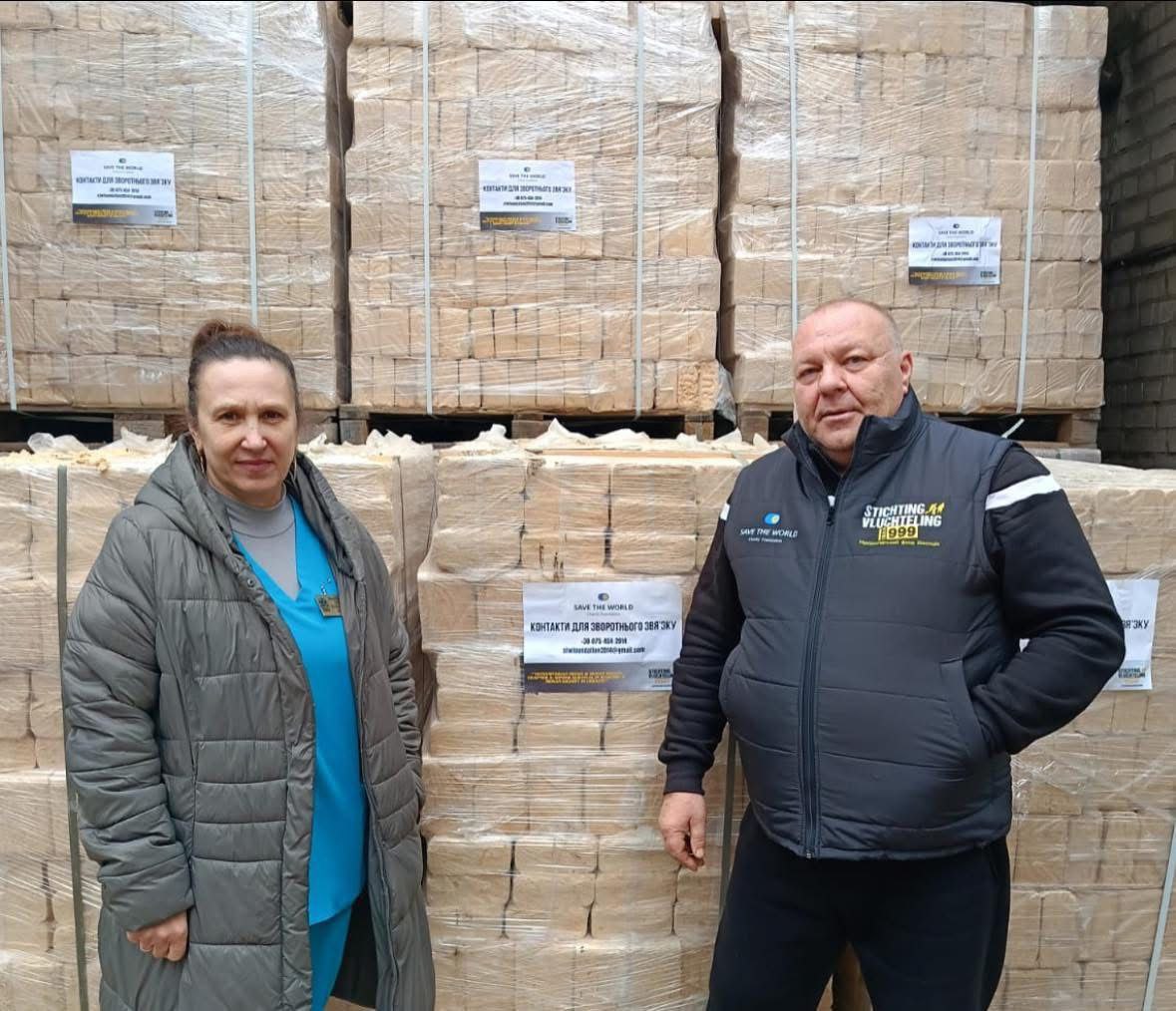 Чоловік та жінка стоять на фоні складених палет із паливними брикетами. На палетах розміщено наклейки благодійного фонду SAVE THE WORLD, а на жилеті чоловіка — логотипи партнерських організацій.