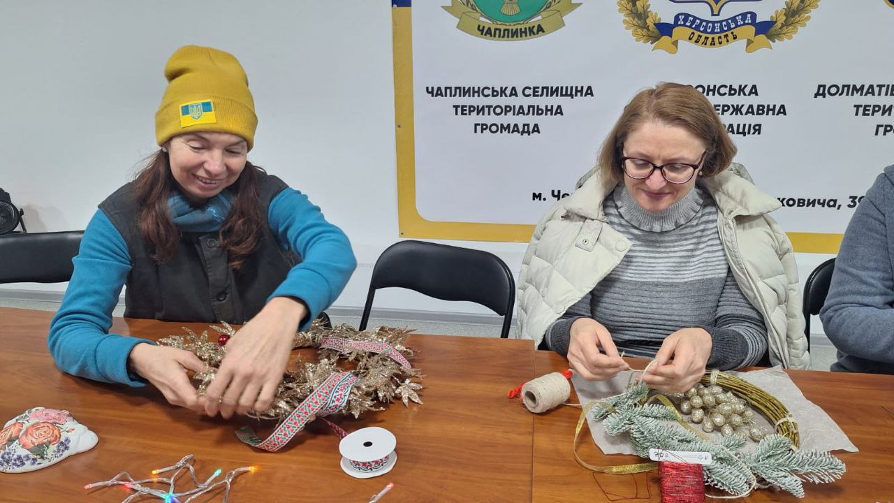 Майстер-клас зі створення різдвяного декору: дві жінки зосереджено працюють над вінками, використовуючи золотисті гілочки, хвою та джутову нитку на фоні банера Чаплинської громади, Херсонської ОДА та Долматівської територіальної громади.