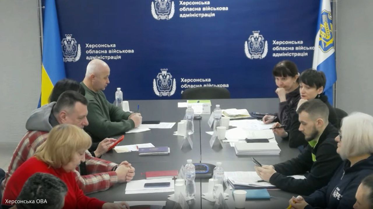 Робоча зустріч у Херсонській обласній військовій адміністрації. Група людей сидить за великим столом під час наради на фоні синього банера з офіційною символікою ОВА та прапорів України.