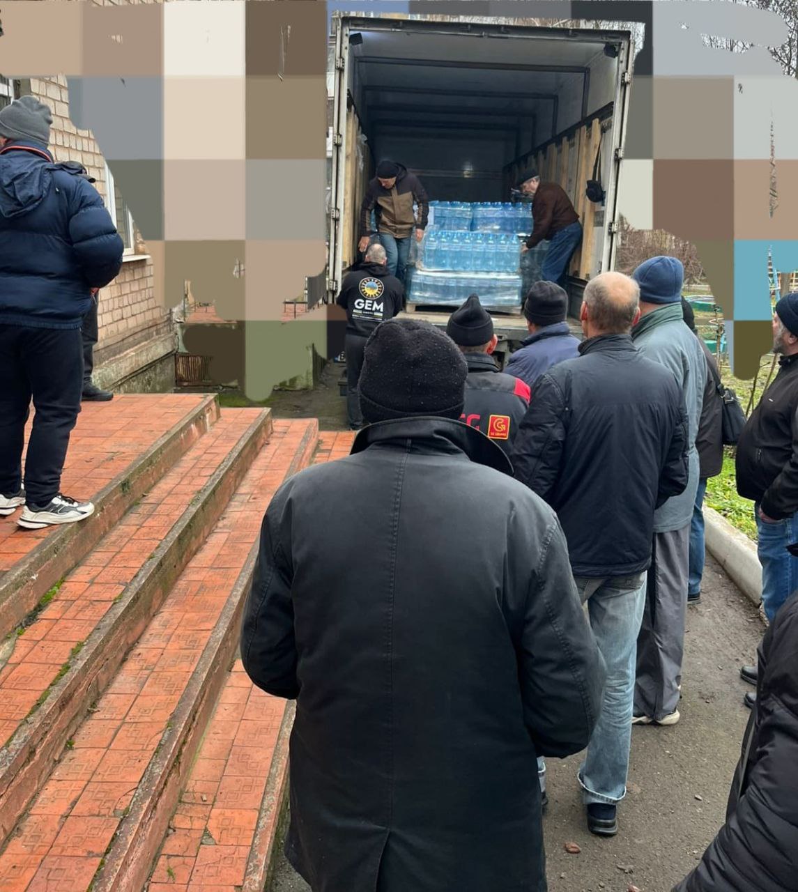 Черга людей біля вантажівки, з якої волонтери розвантажують бутильовану питну воду. Процес відбувається на подвір’ї біля цегляної будівлі.