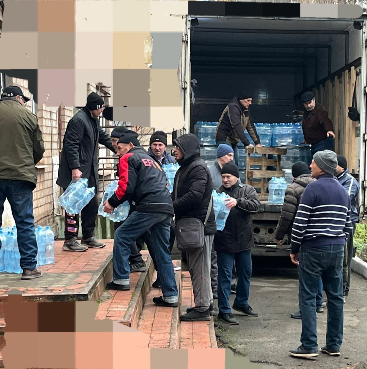 Група чоловіків розвантажує великі бутлі з питною водою з вантажівки. Вони передають воду ланцюжком на цегляний ґанок будівлі.