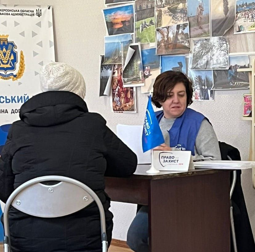 Працівниця гуманітарної організації у синьому жилеті консультує відвідувача за робочим столом. На стіні позаду розміщено колаж із фотографій та інформаційні матеріали.