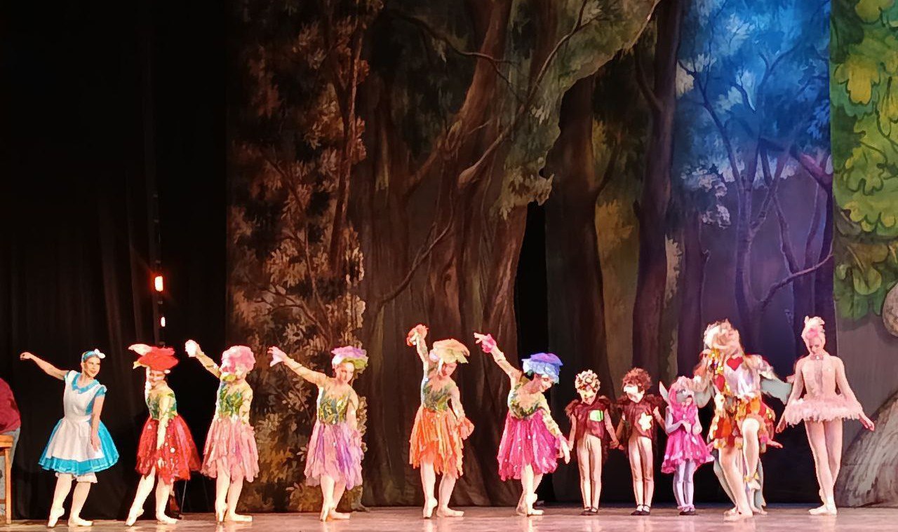 Група артистів балету в яскравих костюмах казкових персонажів позує на сцені театру. Декорації на задньому плані зображують густий чарівний ліс у теплих та насичених кольорах.