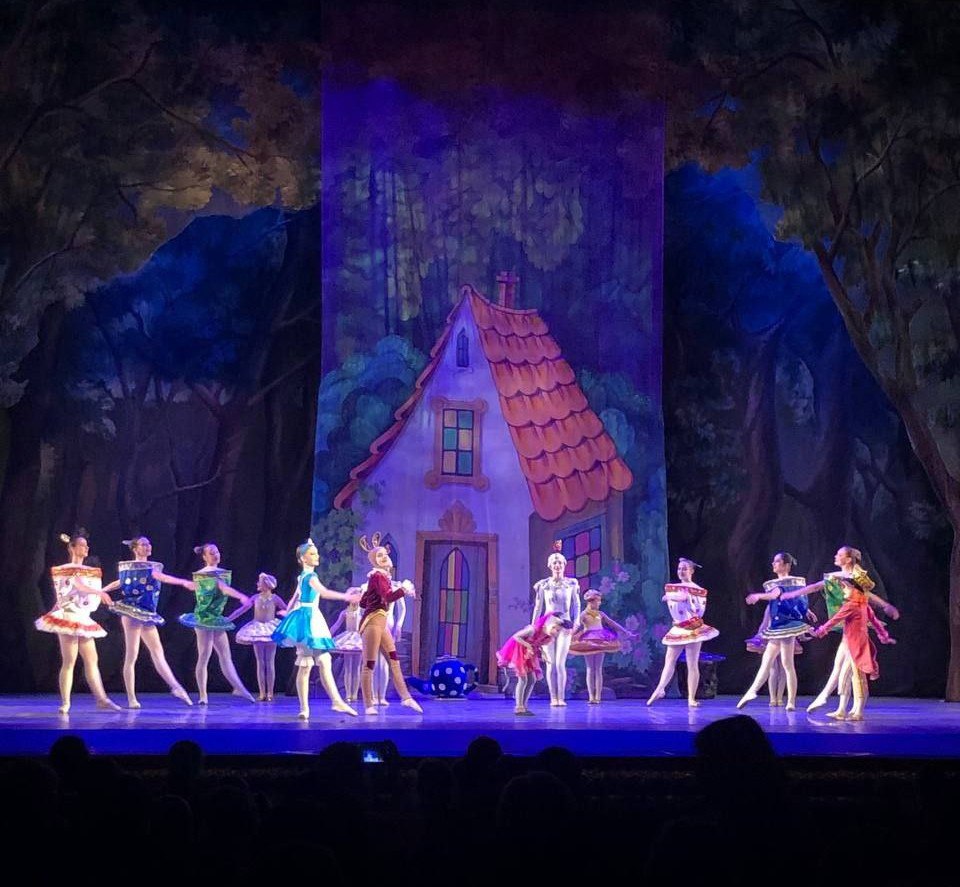 Сценічна постановка балету: група артистів у яскравих різнокольорових костюмах танцює на сцені на тлі декорацій із казковим будиночком та лісом у синіх тонах.