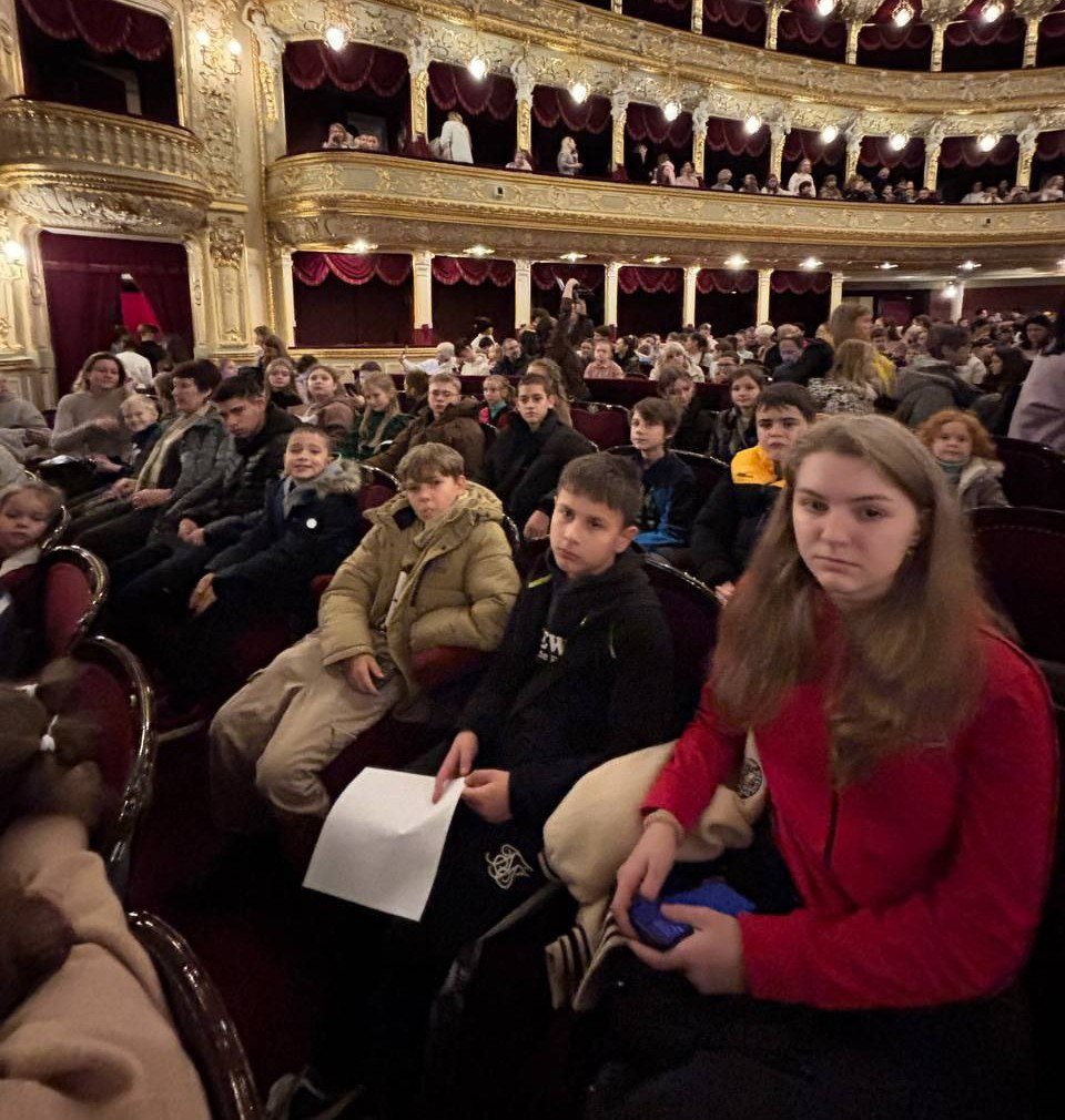 Глядачі, переважно діти та підлітки, сидять у залі театру в очікуванні вистави. Видно класичний інтер'єр з червоними оксамитовими кріслами, ярусами балконів та ліпниною з позолотою.