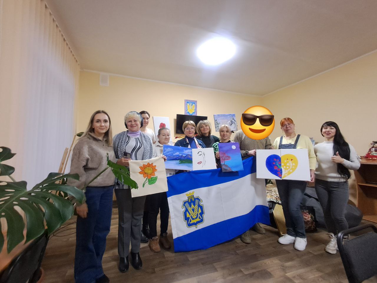 Група жінок стоїть у приміщенні, тримаючи в руках свої творчі роботи: малюнки на полотнах та сумку з розписом. У центрі вони тримають великий синьо-білий прапор із гербом Херсона.