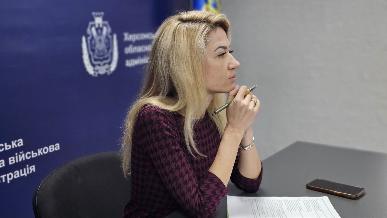 Жінка зі світлим волоссям у темно-бордовій сукні сидить за робочим столом, зосереджено слухаючи співрозмовників. Вона тримає ручку біля підборіддя, перед нею лежать документи, а на фоні видно синій банер із символікою обласної адміністрації.