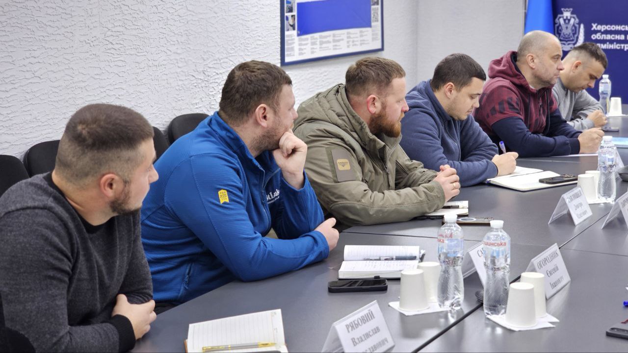 Група чоловіків у повсякденному та тактичному одязі сидить за робочим столом під час наради. Учасники зосереджено слухають, дехто робить нотатки у блокнотах; перед ними на столі стоять пляшки з водою та паперові стакани.