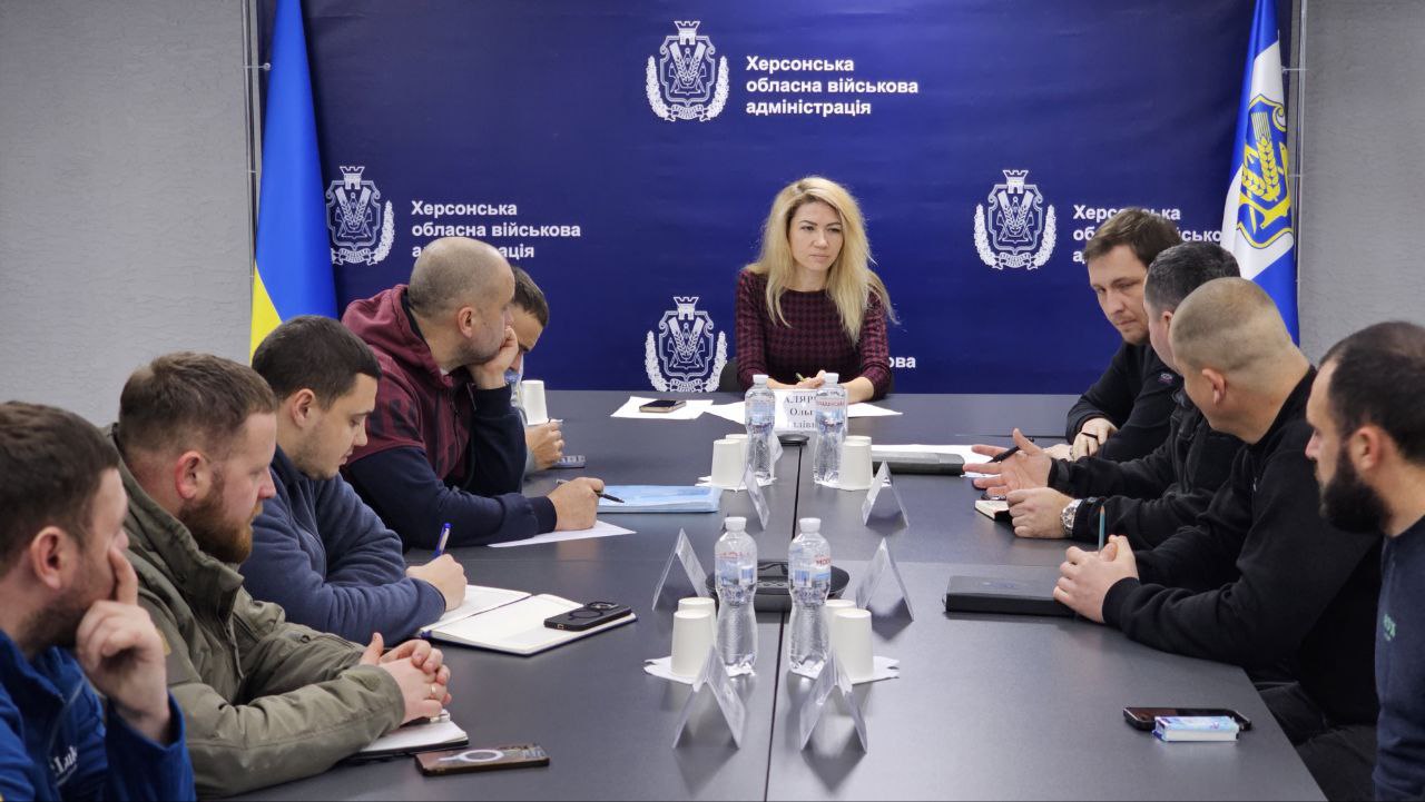 Робоча нарада в офіційному приміщенні: група людей сидить за довгим столом, ведучи обговорення та роблячи нотатки. На фоні розташований синій банер із логотипом Херсонської обласної військової адміністрації та державний прапор України.
