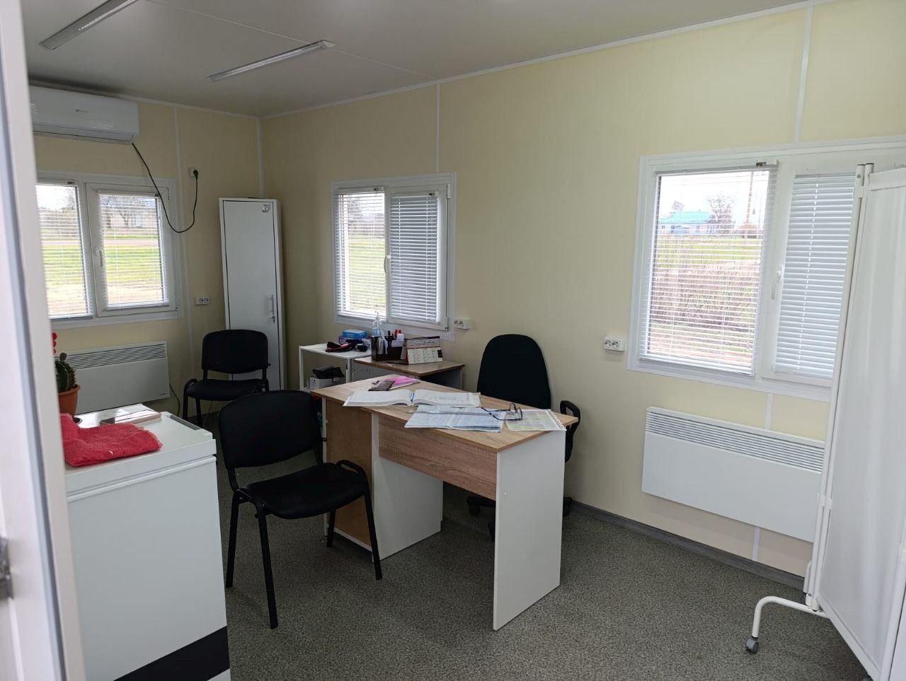 Інтер'єр світлого робочого кабінету або мобільного офісу з бежевими стінами та декількома вікнами з жалюзі. У центрі стоїть письмовий стіл із документами, поруч розташовані офісні стільці, шафа та електричні конвектори для обігріву.