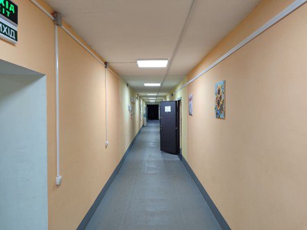 Довгий і чистий коридор у приміщенні з персиковими стінами та сірою підлогою. На стінах висять картини, стеля облаштована вбудованими світильниками, а над дверима ліворуч видно зелене світлове табло ВИХІД.