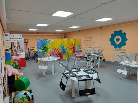 Облаштована дитяча кімната або ігровий центр у приміщенні цивільного захисту. У залі розставлені невеликі столи зі стільцями, стіни прикрашені яскравими малюнками та логотипом центру, а в кутку стоять іграшки, глобус та інформаційний стенд.