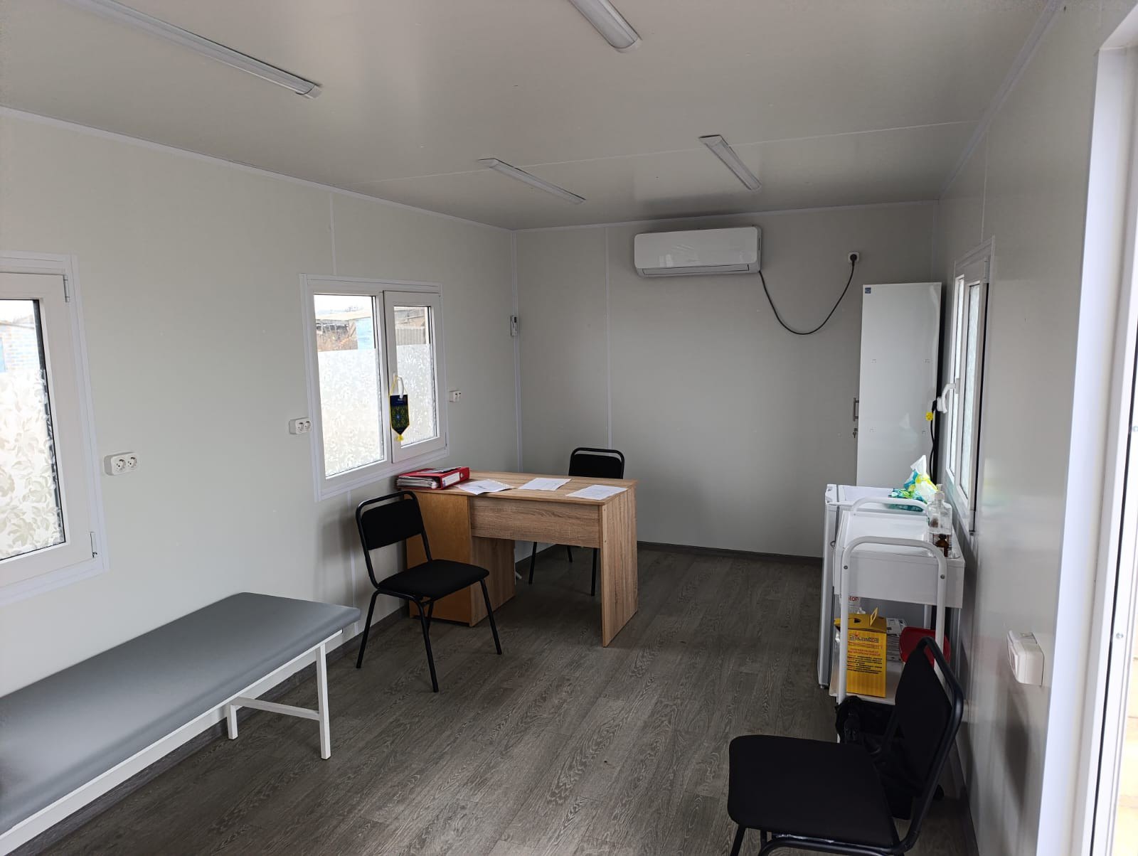 Інтер'єр облаштованого медичного кабінету або пункту допомоги в модульному приміщенні. У кімнаті розташована кушетка, письмовий стіл зі стільцями, медична тумба з антисептиками та кондиціонер на стіні.