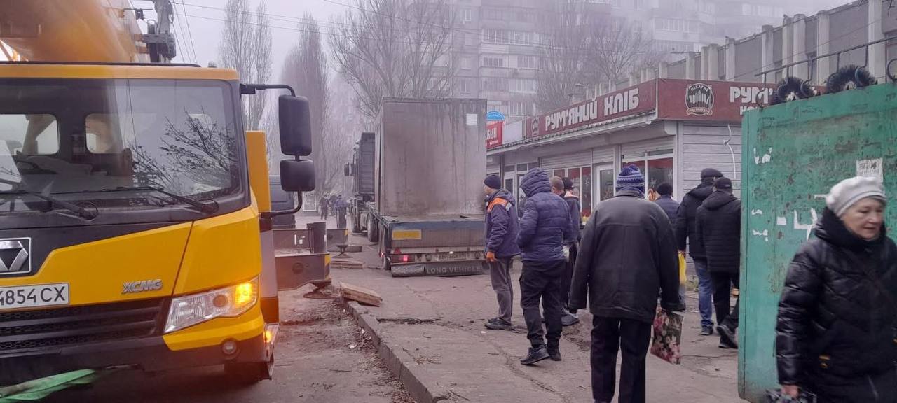 Вулична сцена в туманний день: жовта спецтехніка та вантажівка стоять на узбіччі дороги біля торговельних кіосків. Перехожі у теплому одязі йдуть тротуаром повз припарковані машини.