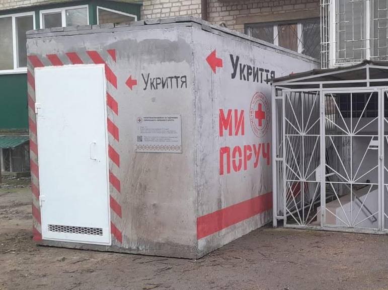 Наземне бетонне укриття модульного типу, встановлене біля житлового будинку. На білій стіні споруди нанесено написи: Укриття зі стрілками, логотип Червоного Хреста та гасло МИ ПОРУЧ великими червоними літерами.