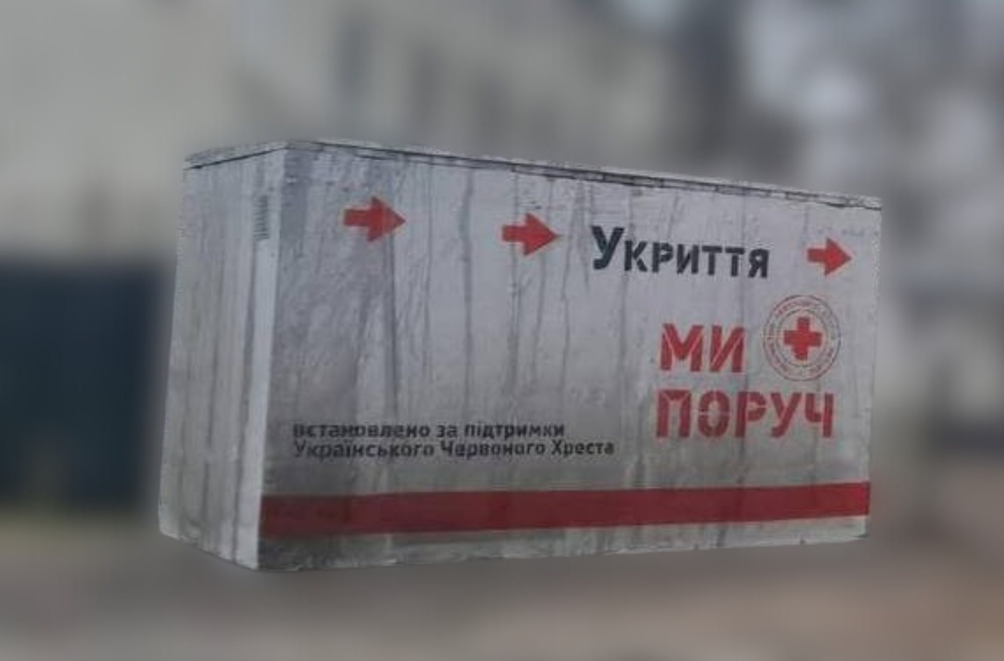 Бетонне мобільне укриття сірого кольору на розмитому фоні міста. На стіні споруди нанесено великий червоний напис: МИ ПОРУЧ, слово Укриття з вказівними стрілками та логотип Червоного Хреста.