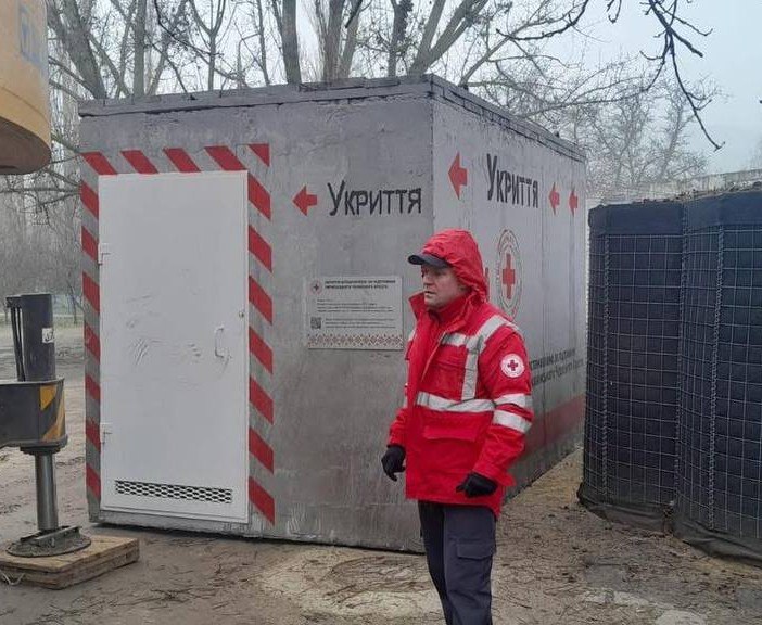 Працівник у червоному форменому одязі з емблемою Червоного Хреста стоїть на вулиці поруч із мобільним бетонним укриттям. На споруді видно написи: Укриття та червоні вказівні стрілки.