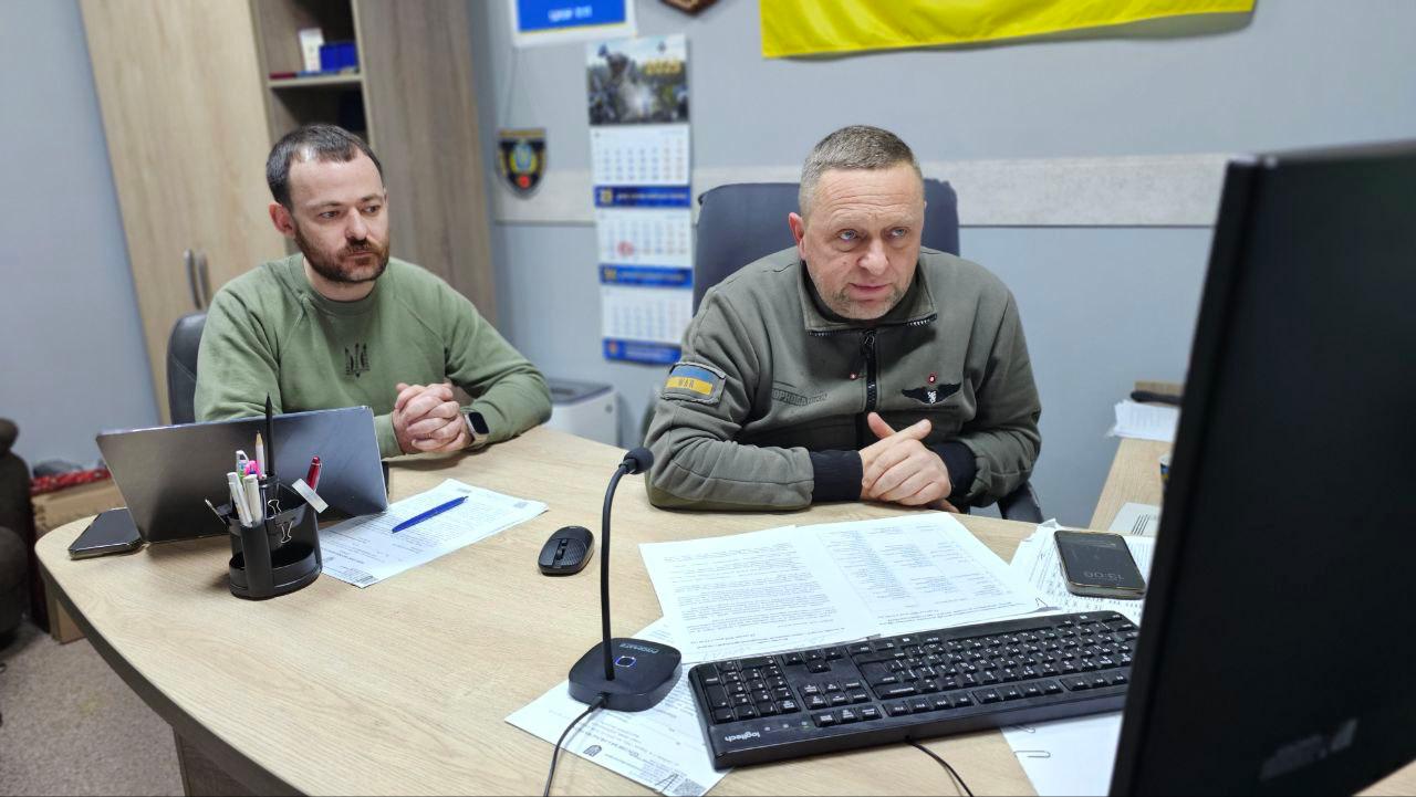 Двоє чоловіків у робочому кабінеті беруть участь в онлайн-нараді, дивлячись на монітор комп'ютера. На столі перед ними розкладені документи, стоїть клавіатура та ноутбук, а на стіні позаду видно календар і жовтий прапор.