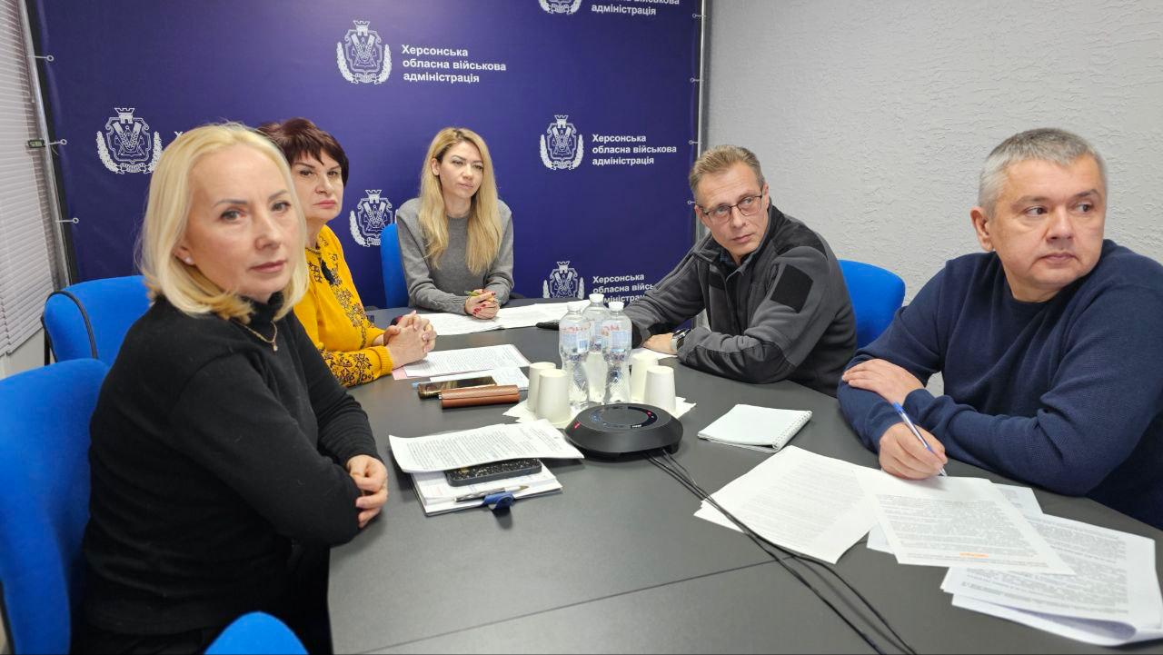 Група людей сидить за робочим столом під час наради на фоні синього банера з логотипом Херсонської обласної військової адміністрації. Учасники обговорення вивчають документи, перед ними стоять пляшки з водою та розкладені записники.